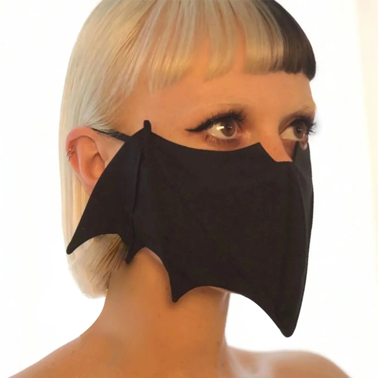 A person with platinum blonde hair and blunt bangs wears a Gay Skate black bat reusable cloth mask, its wings elegantly covering their nose and mouth. The softly lit, blurred background highlights this unique accessory.