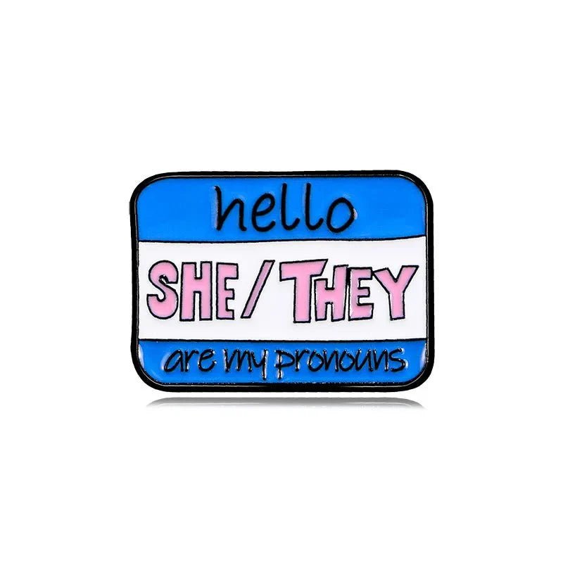 Pronoun pin is shaped like a name tag with blue, white, and pink stripes. It reads: hello SHE/THEY are my pronouns. A colorful accessory—now available for worldwide shipping!.