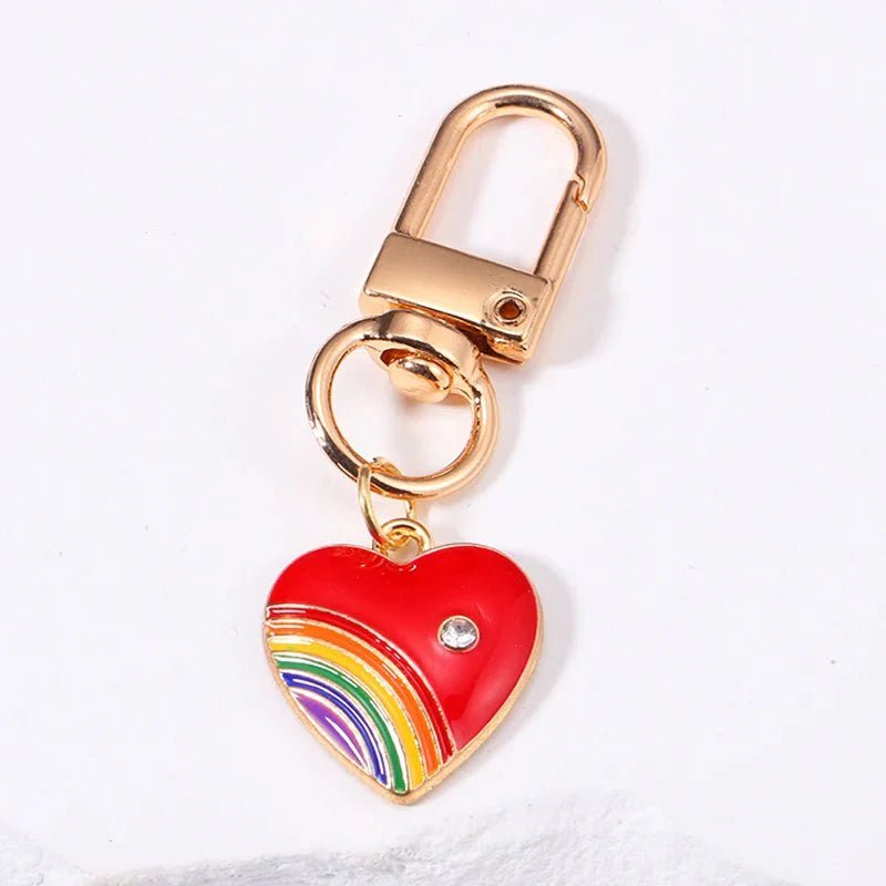 Pride flag keychain with a gold keyring, red heart-shaped charm with a small gem, and a rainbow charm on the lower left. The perfect pride accessory, shown on a crisp white background.