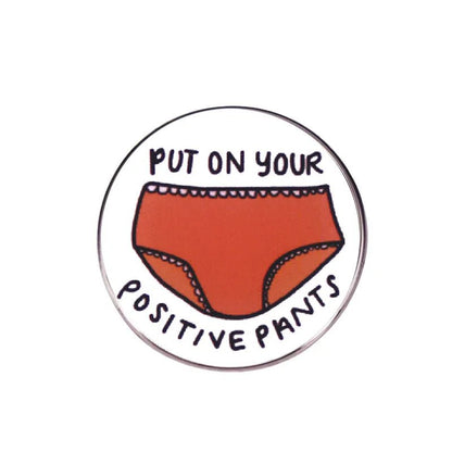 A round enamel pin by My Cutesy Era showcasing red underwear and the phrase PUT ON YOUR POSITIVE PANTS around the illustration.