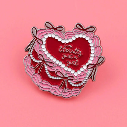 A dreamy heart-shaped enamel badge with pink ruffles, bows, and pearls on a pink background. Features Im Literally Just a Girl in white cursive—perfect for fans of pink and red cake badge designs.