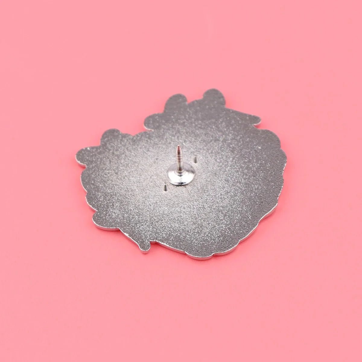 metallic enamel badge with an irregular shape is face down on a pink background, revealing its sharp post and backing attachment.