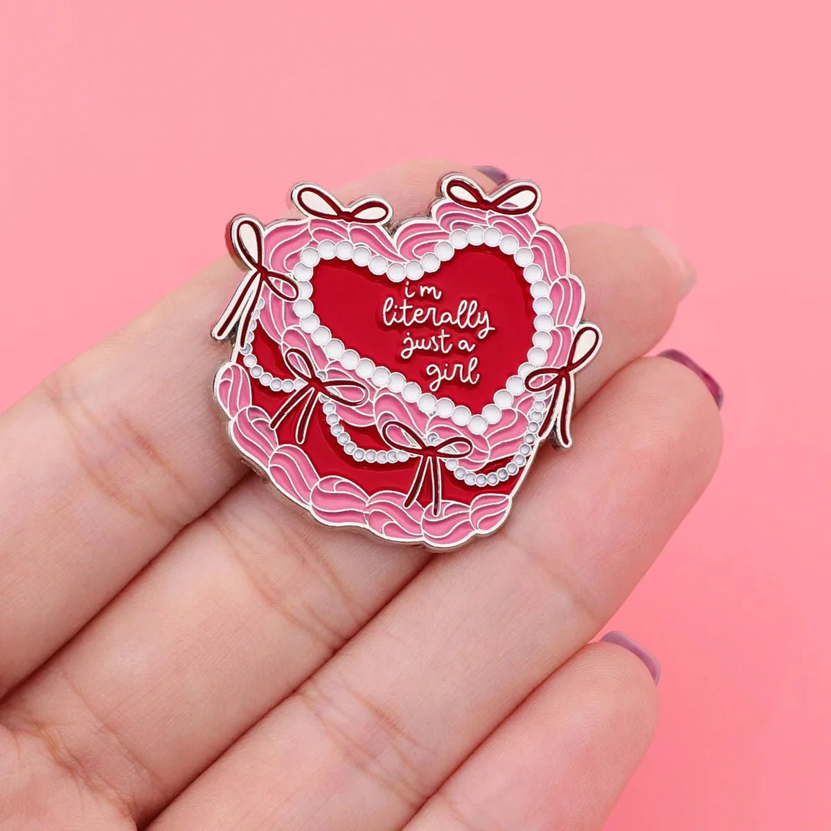 A hand holds a heart-shaped enamel badge styled as a pink and red cake with icing, pearls, bows, and the cursive phrase Im Literally Just a Girl on a red background against pink.