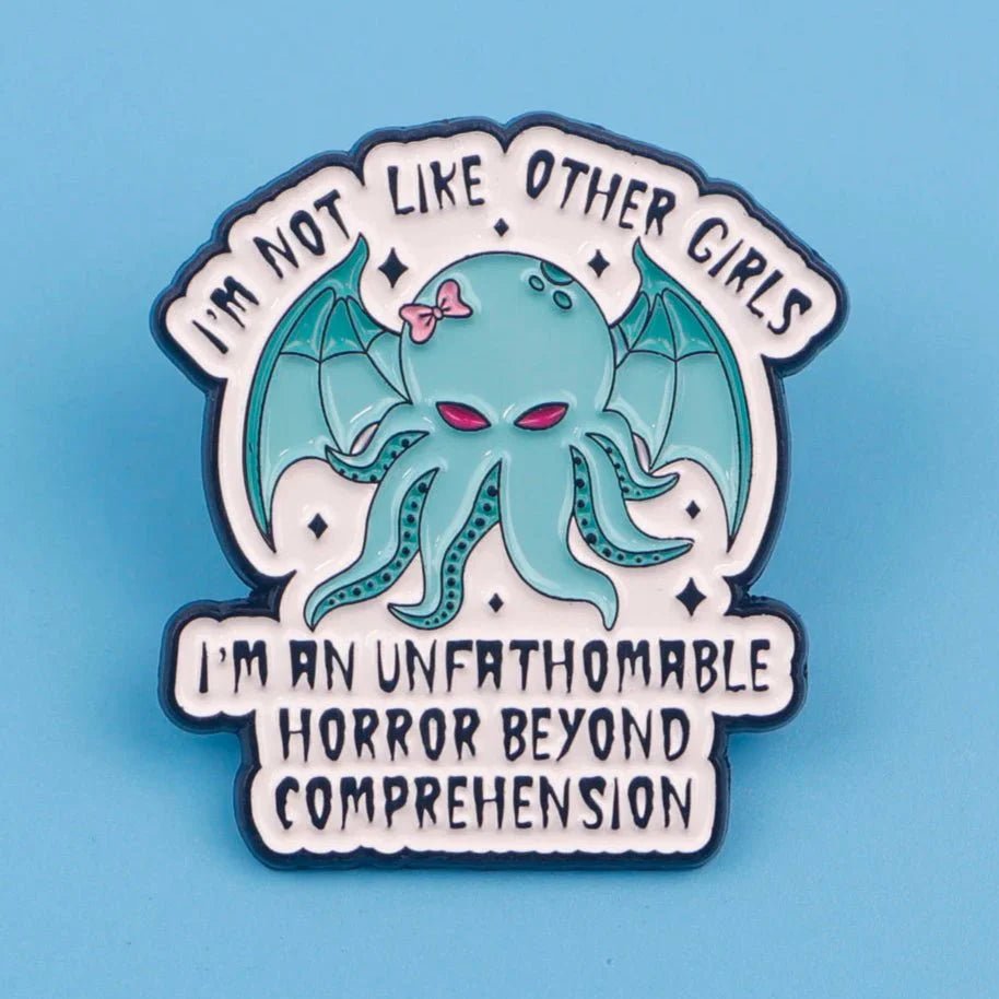 The Kiss Me Later enamel badge features a cute, cartoon Cthulhu with a pink bow and bat wings, surrounded by the text: I’m not like other girls. I’m an unfathomable horror beyond comprehension, on a light blue background.