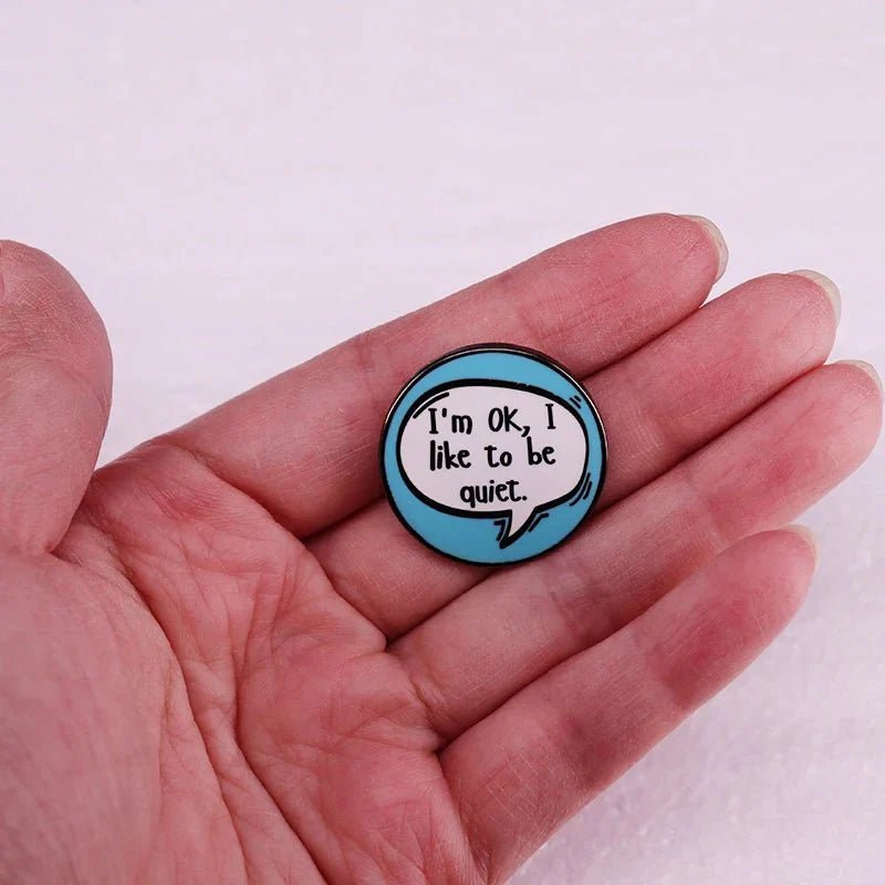 A hand holds a Gay Skate Introvert Enamel Pin—a round blue speech bubble reading Im OK, I like to be quiet—ideal for anyone seeking unique pride accessories.
