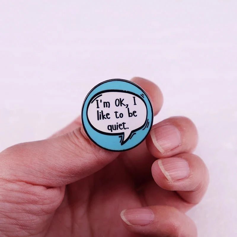A hand holds a Gay Skate Introvert Enamel Pin with a blue speech bubble saying, Im OK, I like to be quiet—a perfect pride accessory for anyone expressing their personality.