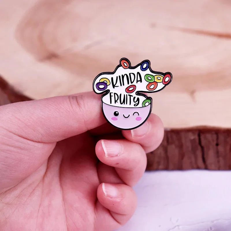 A hand holds a Kiss Me Later enamel pin shaped like a smiling bowl with colorful cereal rings and “KINDA FRUITY” above—perfect for showing off your playful and queer pride!.