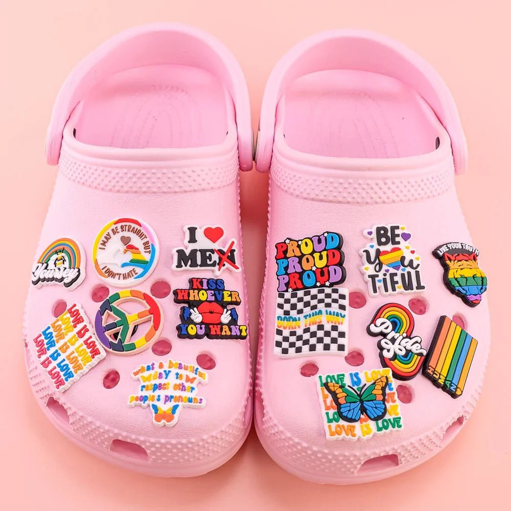 A pair of crocs adorned with vibrant pride charms and pins—like the Rainbow Flag and uplifting phrases—is showcased on a matching pink background.