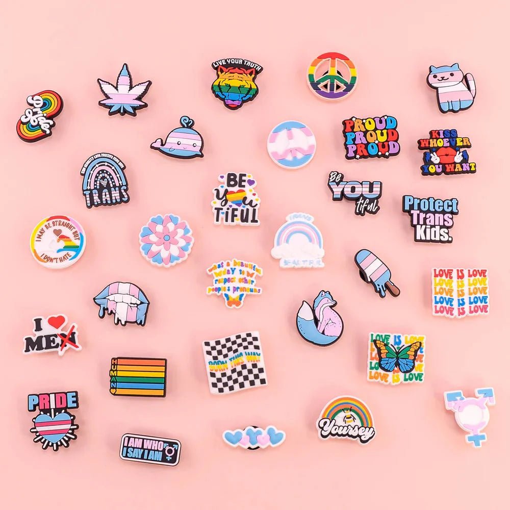 colorful LGBTQ+ and pride pins, featuring hearts, “Be You tiful” slogans, the Trans Flag, Rainbow Flag, and other pride flags—all beautifully arranged on a light pink background.