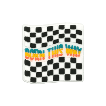 A wavy black-and-white checkered flag by Kiss Me Later, featuring BORN THIS WAY in blue, yellow, and red across the center, accented with pride-themed charms.