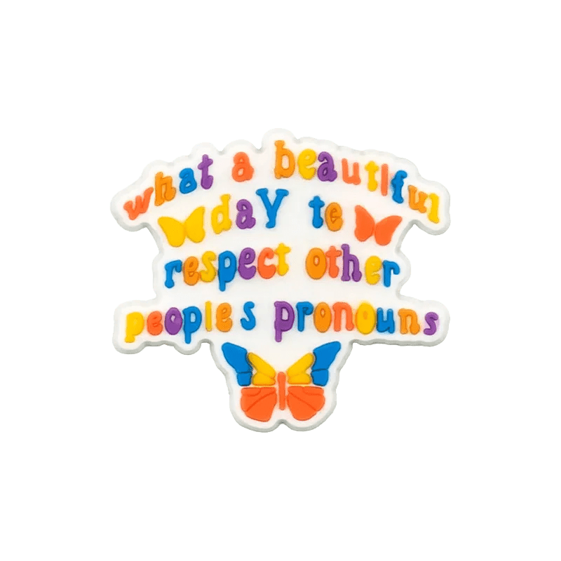 Kiss Me Later’s design features vibrant, colorful text with three butterflies and subtle Rainbow and Trans Flag motifs that say: “What a beautiful day to respect other peoples pronouns.”.