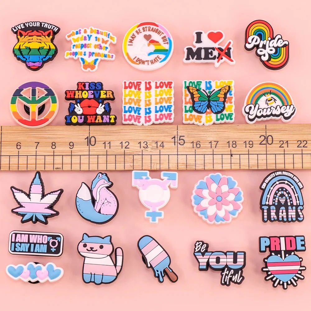 A vibrant selection of LGBTQ+ and pride-themed pins, including charms and the Rainbow Flag, are displayed on pink with a wooden ruler for scale. Designs feature rainbows, hearts, flags, animals, and positive affirmations.
