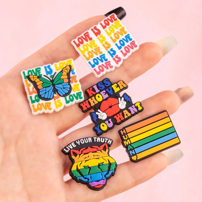 A hand holds five Kiss Me Later pride charms with LGBTQ+ messages: “LOVE IS LOVE,” a butterfly, the Rainbow Flag, “KISS WHOEVER YOU WANT,” a rainbow-striped “HUMAN,” and a tiger with “LIVE YOUR TRUTH.”.