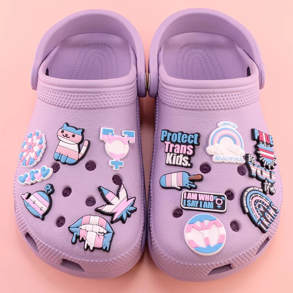 A pair of Kiss Me Later pastel purple Crocs feature pride charms with Trans Flag colors, messages like Protect Trans Kids, a unicorn, rainbows, and Rainbow Flag designs, set against a pink background.