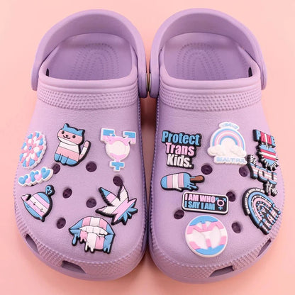 A pair of Kiss Me Later pastel purple Crocs feature pride charms with Trans Flag colors, messages like Protect Trans Kids, a unicorn, rainbows, and Rainbow Flag designs, set against a pink background.