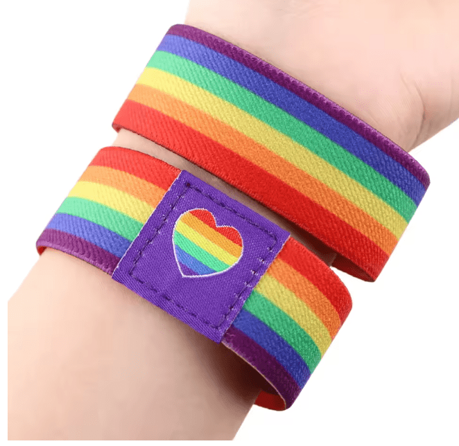 A close-up of a wrist wearing two rainbow-striped Pride wristbands from Gay Skate; one has a purple patch with a rainbow heart, representing LGBTQ+ pride and the rainbow flag’s spirit.
