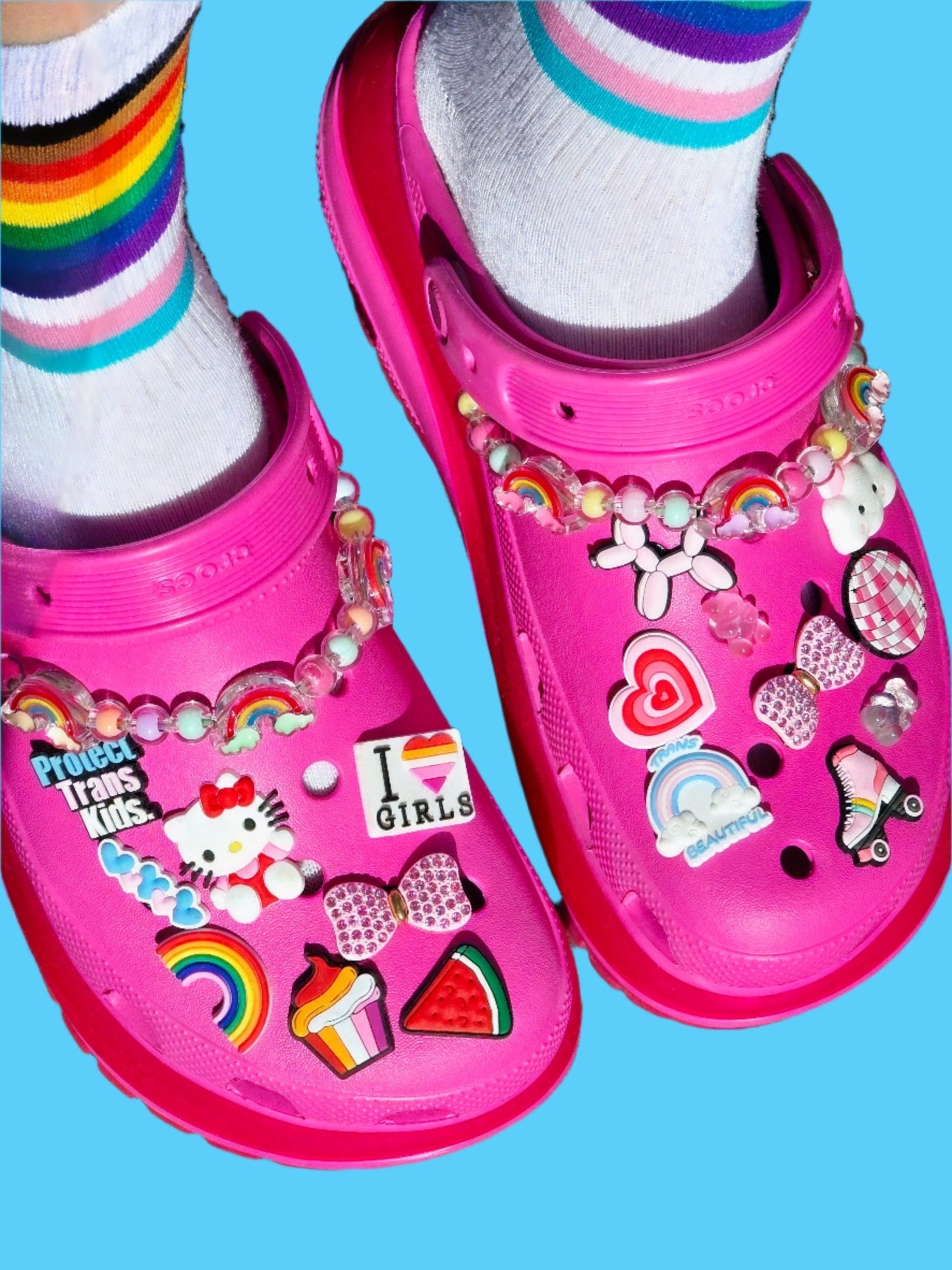Gay Skate bright pink Crocs adorned with colorful pins and pride charms—rainbows, hearts, Hello Kitty, and the Trans Flag—paired with white socks featuring rainbow stripes, all set on a blue background.