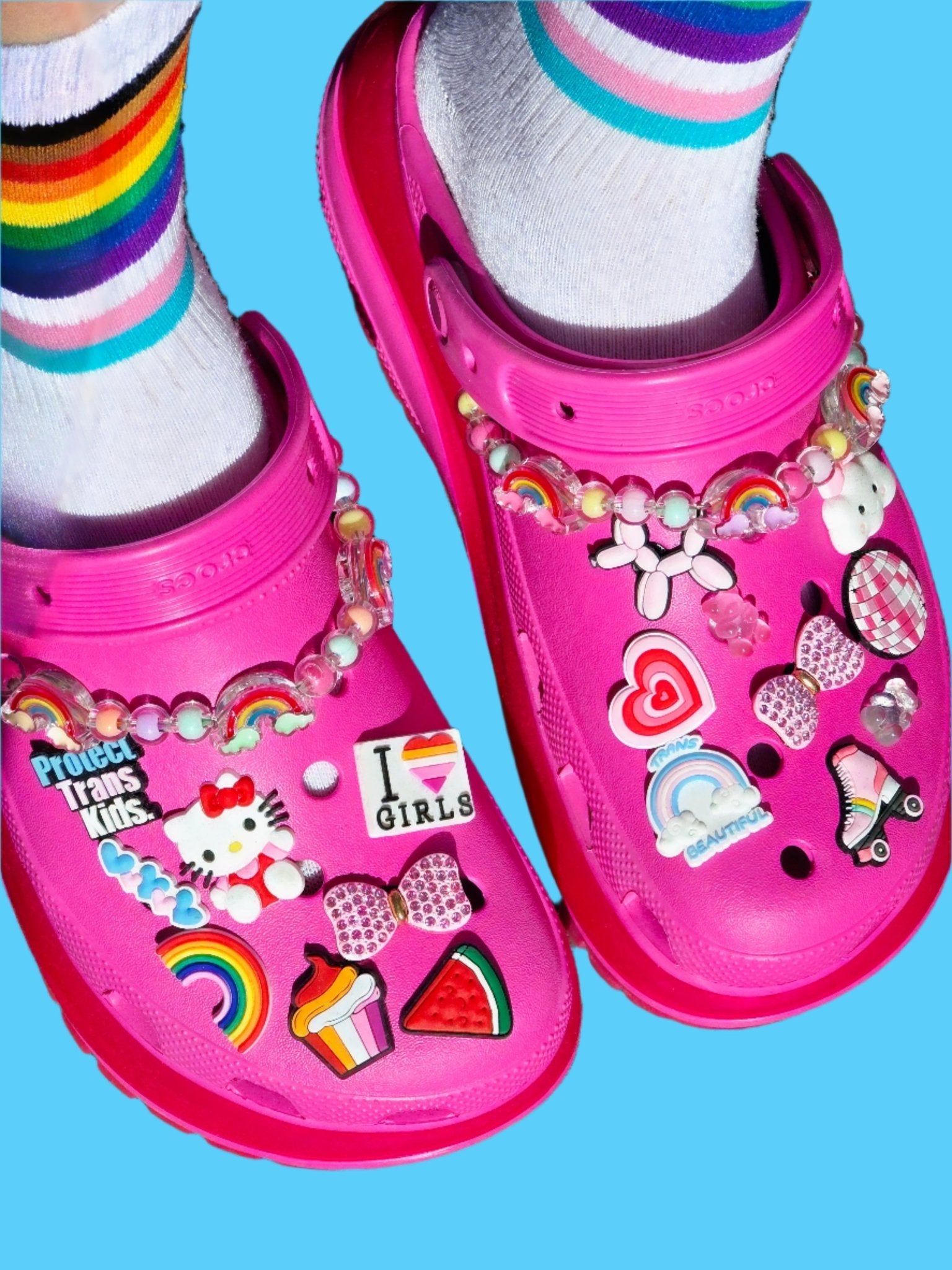 Gay Skate bright pink Crocs adorned with colorful pins and pride charms—rainbows, hearts, Hello Kitty, and the Trans Flag—paired with white socks featuring rainbow stripes, all set on a blue background.