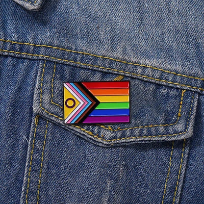 A rectangular Progress Pride rainbow pin by Gay Skate, featuring rainbow stripes and a chevron with black, brown, blue, pink, white, yellow, and a purple circle for inclusivity, is attached to the pocket of a blue denim jacket.