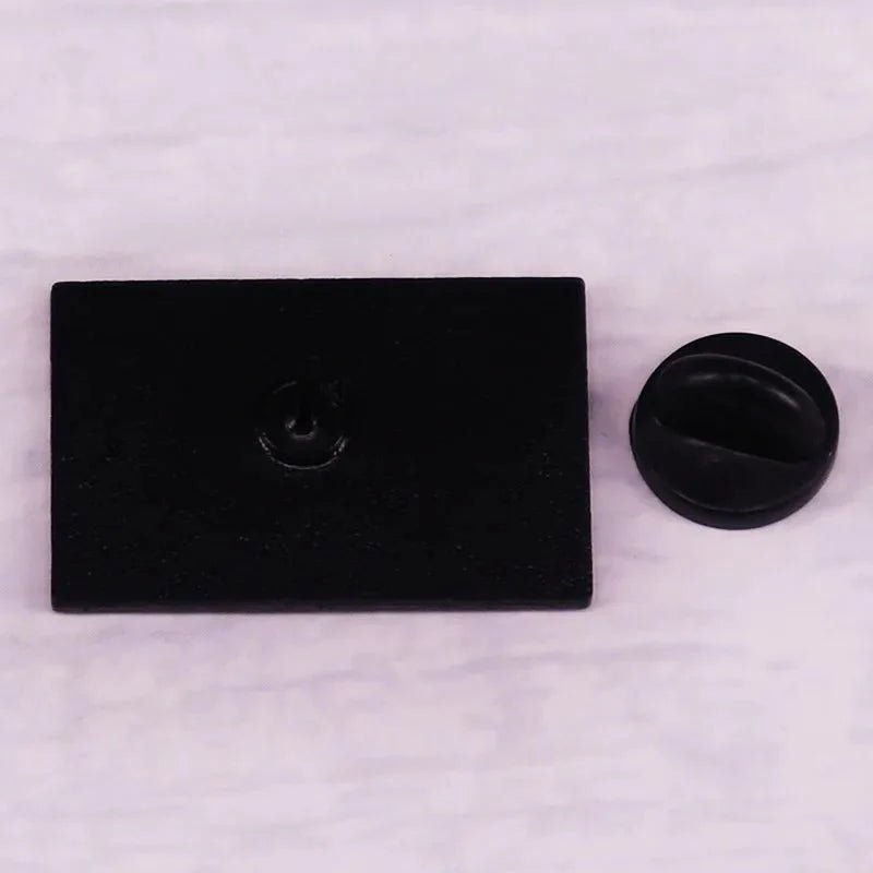 A rectangular black Gay Skate pin backing plate sits beside a round black pin clasp—ideal for securing your favorite inclusive pride items—on a light, textured surface.