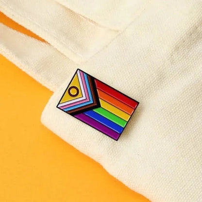 The Gay Skate Progress Pride flag pin features rainbow stripes and pink, blue, white, brown, and black chevrons. Resting on cream fabric with a yellow backdrop, this inclusive pride accessory celebrates diversity.