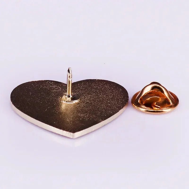 A metallic heart-shaped pin inspired by the Progress pride flag, features a pointed back and separate round gold clasp, both displayed on a white surface.