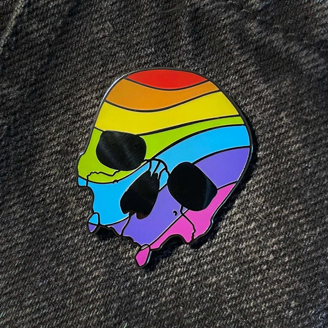 rainbow pride skull enamel pin with vibrant wavy stripes is shown on dark textured fabric. Express yourself with this unique accessory—worldwide shipping available.
