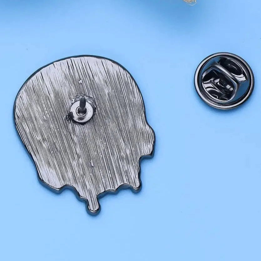 Rainbow Pride Skull enamel pin shaped like a melting skull rests on a blue surface with its detached clasp nearby; part of a woven item is at the top left. Enjoy worldwide shipping.