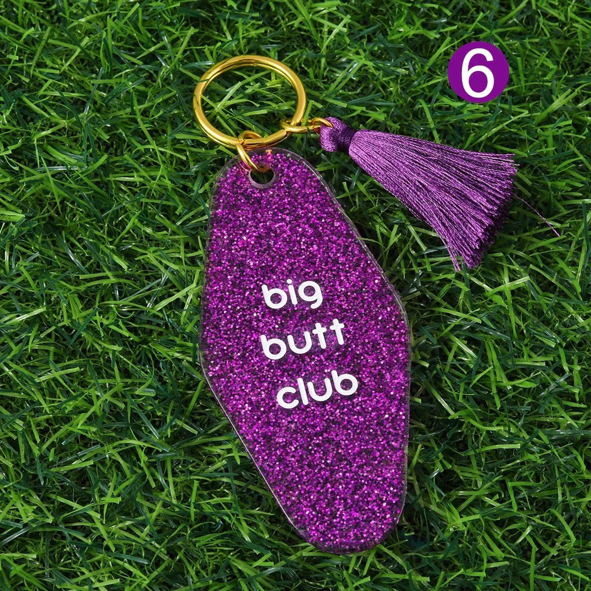 A purple glitter Big Butt Club hotel key ring with a matching tassel rests on green grass. A purple circle with the number 6 is in the top right. Worldwide shipping available.