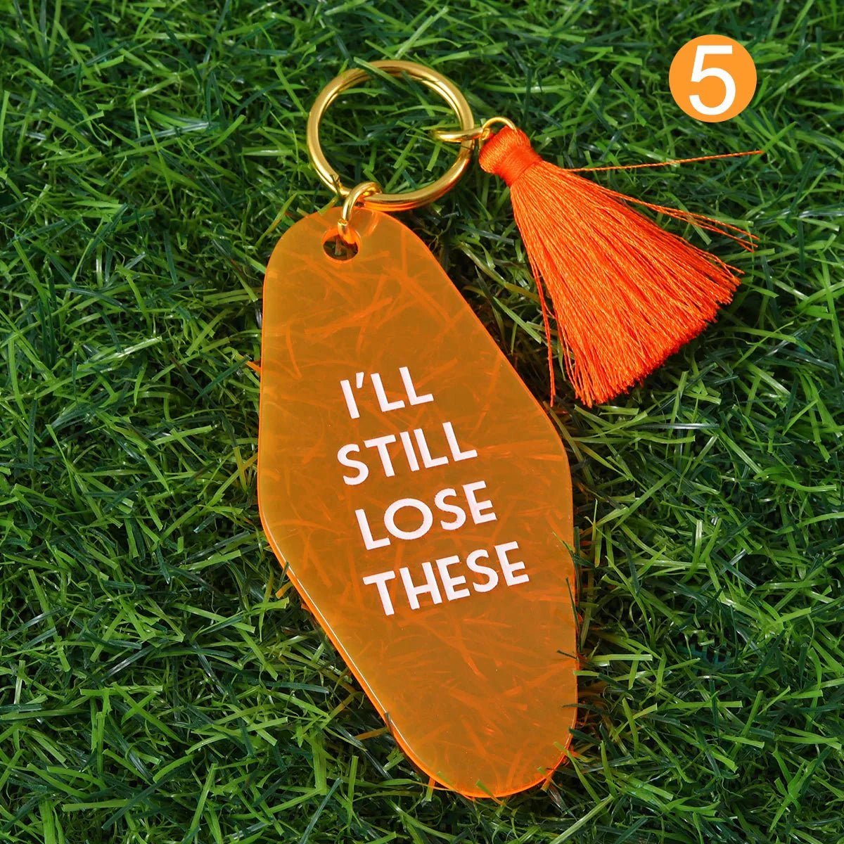 An orange kitsch hotel key ring with white “I’LL STILL LOSE THESE” text, an orange tassel, and a gold 12cm ring rests on green grass. An orange circle with the number 5 is in the top right corner.
