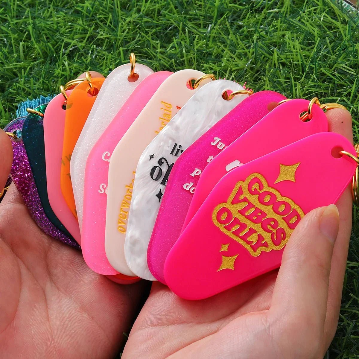 A hand fans out several colorful, retro hotel key rings (12cm), each with unique phrases and hues. The front reads GOOD VIBES ONLY in gold, set against green grass. Worldwide shipping available.