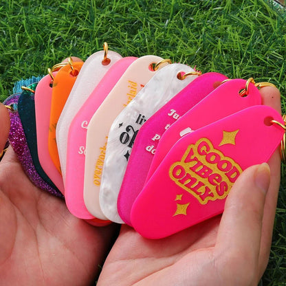 A hand fans out several colorful, retro hotel key rings (12cm), each with unique phrases and hues. The front reads GOOD VIBES ONLY in gold, set against green grass. Worldwide shipping available.