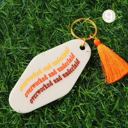 hotel key ring with an orange tassel lies on grass. The 12cm keychain reads overworked and underlaid in yellow, orange, and red text, plus a white number 9. Worldwide shipping available.