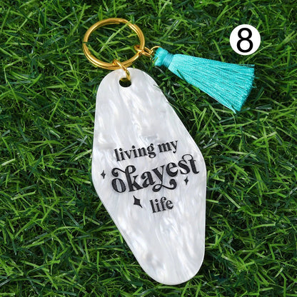 A white marbled hotel key ring with black living my okayest life text, gold 12cm ring, turquoise tassel; number 8 at top right. Photo on green grass. Ships worldwide.