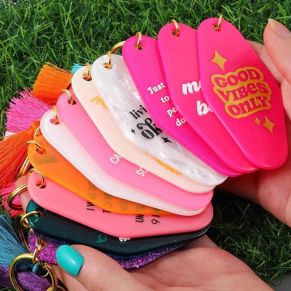 A hand holds colorful hotel key rings with motivational phrases like a bright pink GOOD VIBES ONLY, fanned out against a grassy background. Worldwide shipping available.