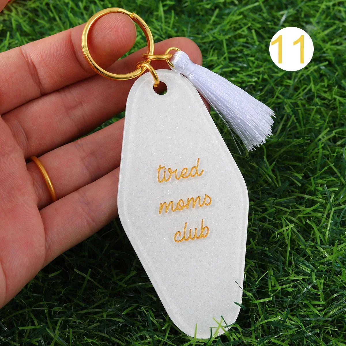 A hand holds a white Tired Moms Club hotel key ring featuring gold text, a gold ring, and white tassel over green grass. A yellow 11 is in the top right. Worldwide shipping available.