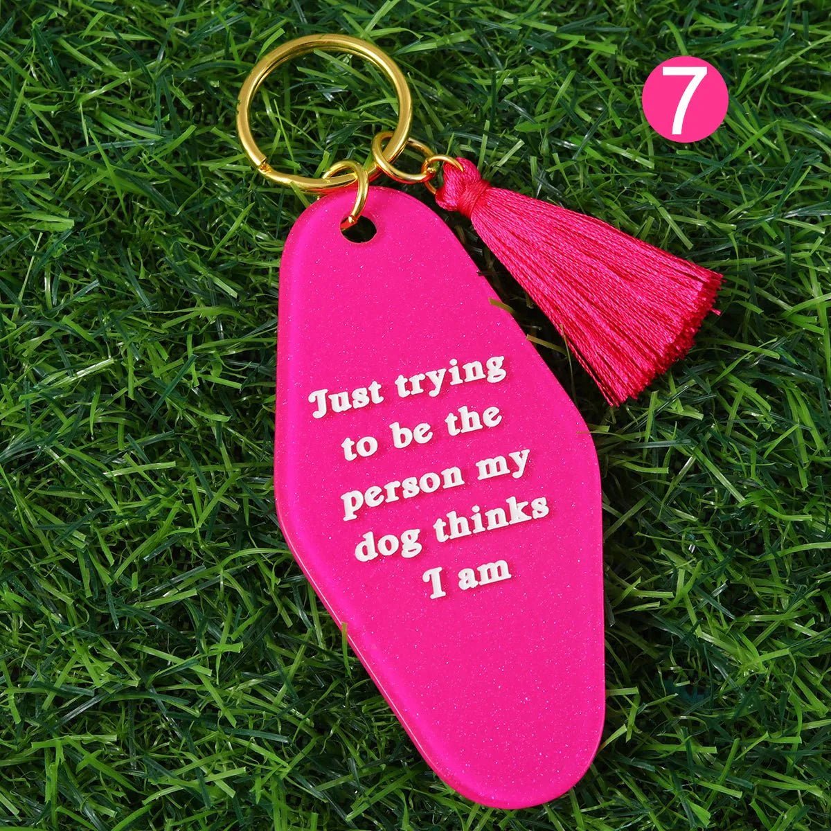 A Kiss Me Later pink kitsch hotel key ring with tassel lies on grass. The keychain says, Just trying to be the person my dog thinks I am. A pink circle with the number 7 is in the top right. Ships worldwide.
