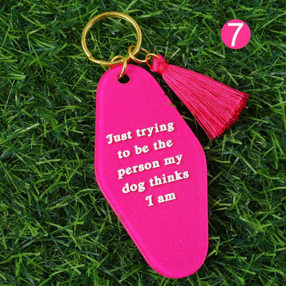 A Kiss Me Later pink kitsch hotel key ring with tassel lies on grass. The keychain says, Just trying to be the person my dog thinks I am. A pink circle with the number 7 is in the top right. Ships worldwide.