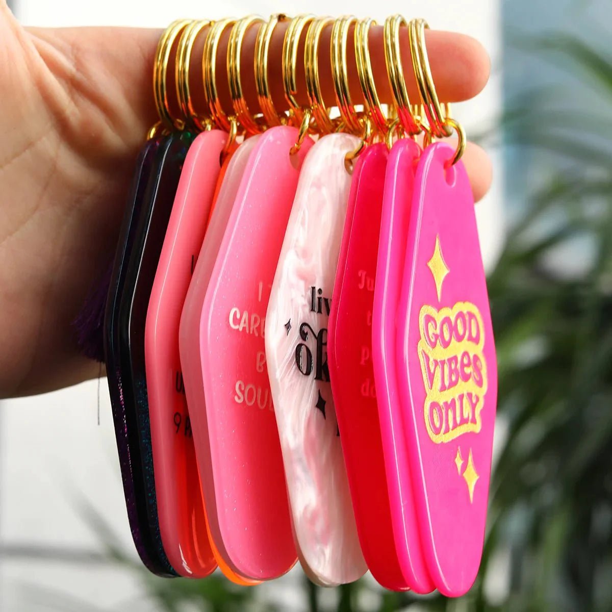 A hand holds several colorful, coffin-shaped hotel key rings with gold rings. One bright pink, 12cm key ring reads “GOOD VIBES ONLY” in yellow letters; other unique designs have visible text.