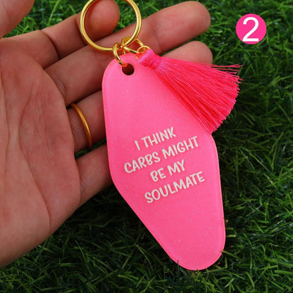 A hand holds a bright pink  hotel key ring (12cm) with a tassel and “I THINK CARBS MIGHT BE MY SOULMATE” in white. The key ring pops against green grass, with a pink circle featuring the number 2 in the corner.