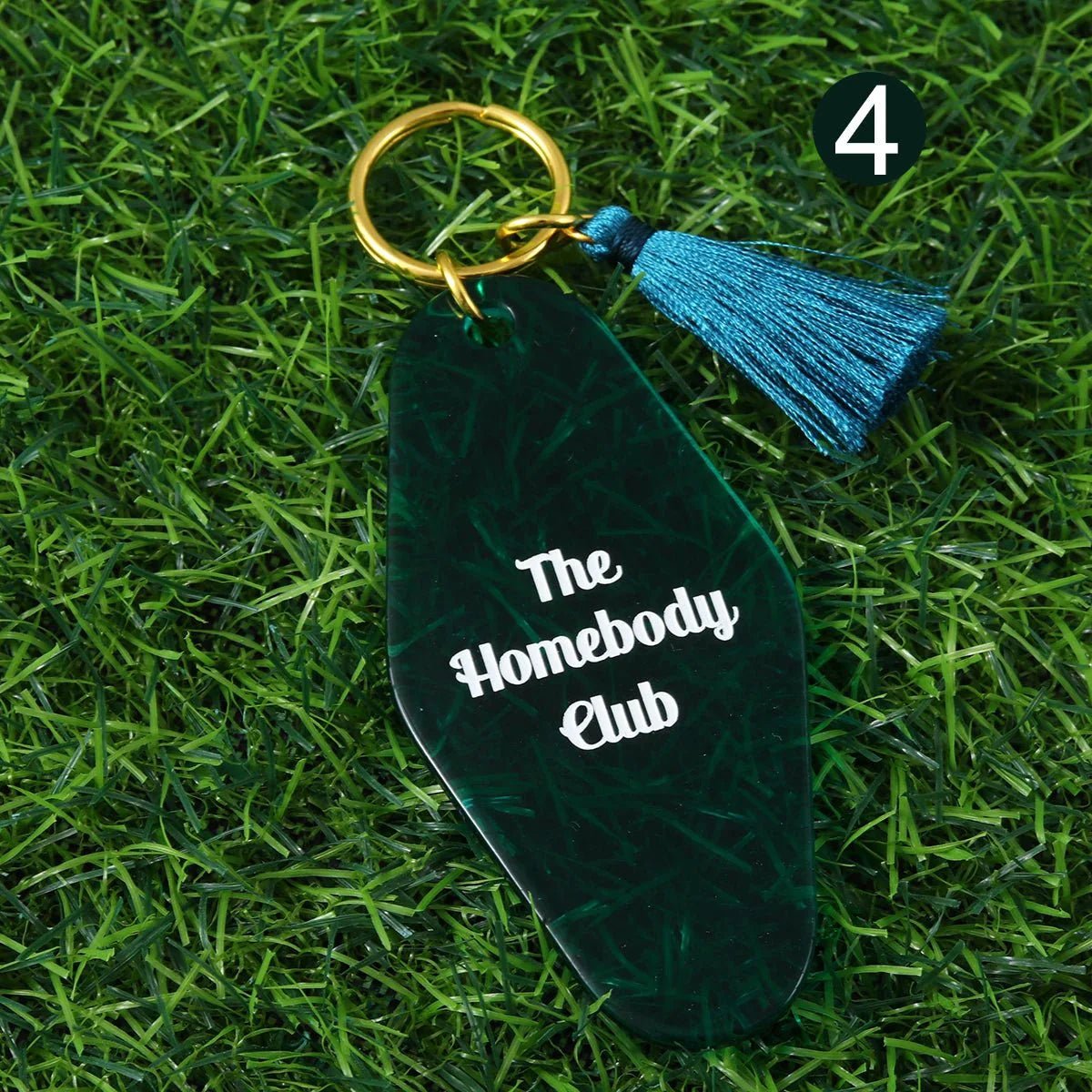 A dark green hotel key ring by  features The Homebody Club in white, a gold keyring, and teal tassel. It lies on artificial grass with a white number 4 in a dark circle. Ships worldwide.
