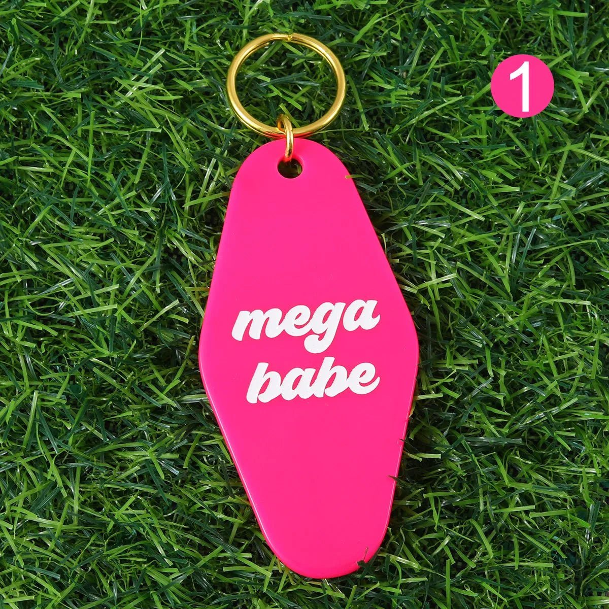 A bright pink mega babe hotel key ring with white text and a 12cm gold ring lies on grass. A pink circle with the number 1 is in the corner. Worldwide shipping available.