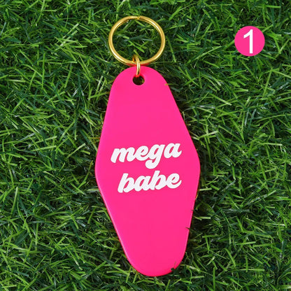 A bright pink mega babe hotel key ring with white text and a 12cm gold ring lies on grass. A pink circle with the number 1 is in the corner. Worldwide shipping available.