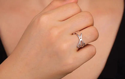 A hand wearing a Stainless Steel Scissor Ring on the middle finger, paired with subtle sapphic pride accessories. The background is blurred, and the person is dressed in a black top.