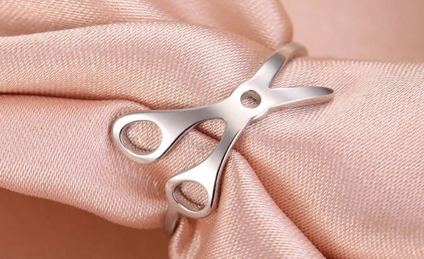 A stainless steel Scissor Ring shaped like scissors, rests on smooth light pink satin—a chic accessory celebrating sapphic pride.