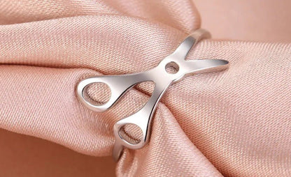 A stainless steel Scissor Ring shaped like scissors, rests on smooth light pink satin—a chic accessory celebrating sapphic pride.