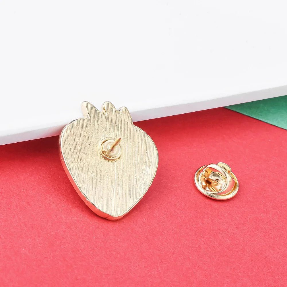 A gold Strawberry Vulva pin inspired by feminist and sapphic WLW themes, sits on a red surface with its backing nearby. A white card and green background highlight the vibrant display.