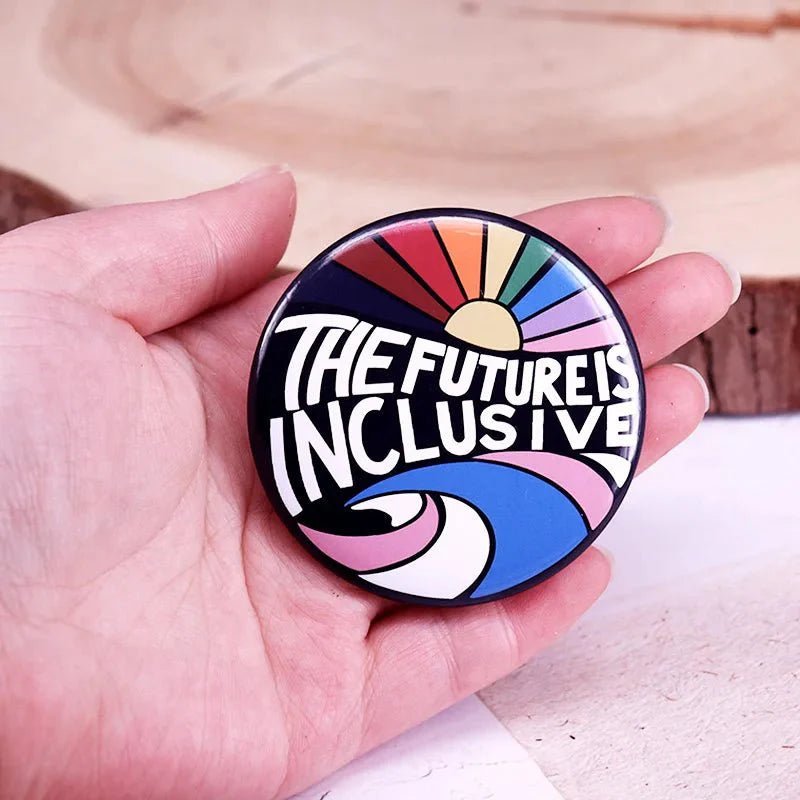 A hand holds a round Gay Skate button badge featuring a colorful design with the words The Future Is Inclusive set against a rainbow pride, sun, and wave background.