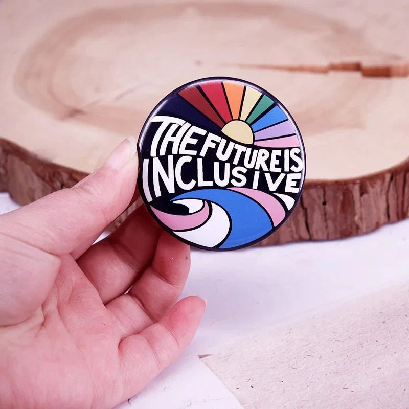 A hand holds a round Gay Skate button badge featuring bold The future is inclusive text with vibrant abstract rainbow designs, set against a wooden surface background.