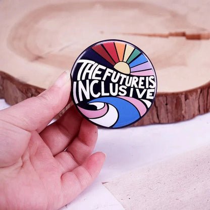 A hand holds a round Gay Skate button badge featuring bold The future is inclusive text with vibrant abstract rainbow designs, set against a wooden surface background.